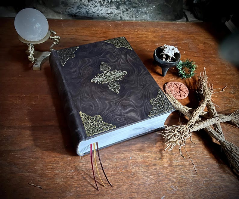 K&ouml;nnte beinhalten: Ein dunkelbraunes, ledergebundenes Buch mit goldenen Verzierungen und einem dekorativen Kreuzmuster auf dem Einband. Das Buch hat drei rote, goldene und wei&szlig;e Lesezeichen aus Band. Das Buch liegt auf einem Holztisch mit anderen Dekorationsgegenst&auml;nden.