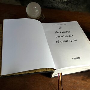 K&ouml;nnte beinhalten: Ein geschlossenes Buch mit einem wei&szlig;en Einband und goldenen Akzenten. Der Titel auf dem Einband lautet "The Element Encyclopedia of 5000 Spells" von Judith Ellen.