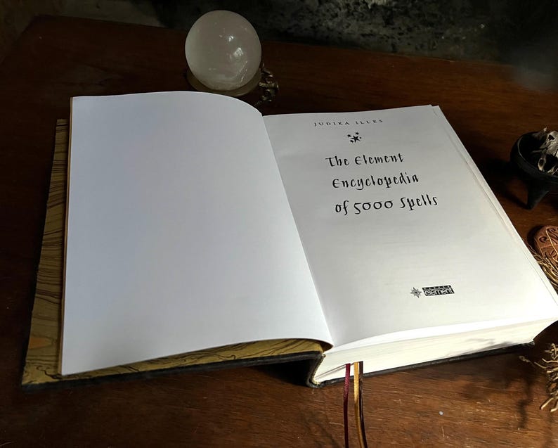 K&ouml;nnte beinhalten: Ein geschlossenes Buch mit einem wei&szlig;en Einband und goldenen Akzenten. Der Titel auf dem Einband lautet "The Element Encyclopedia of 5000 Spells" von Judith Ellen.