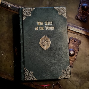 Puede incluir: Un libro encuadernado en cuero verde oscuro con detalles dorados y el t&iacute;tulo "El Se&ntilde;or de los Anillos" en relieve en la portada.