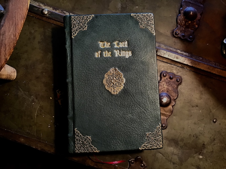 Puede incluir: Un libro encuadernado en cuero verde oscuro con detalles dorados y el t&iacute;tulo "El Se&ntilde;or de los Anillos" en relieve en la portada.