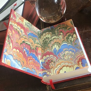Puede incluir: Un diario encuadernado en cuero rojo con papeles de marfil marmoleados. El marmoleado presenta un patr&oacute;n de remolino de rojo, azul, verde, amarillo y blanco.