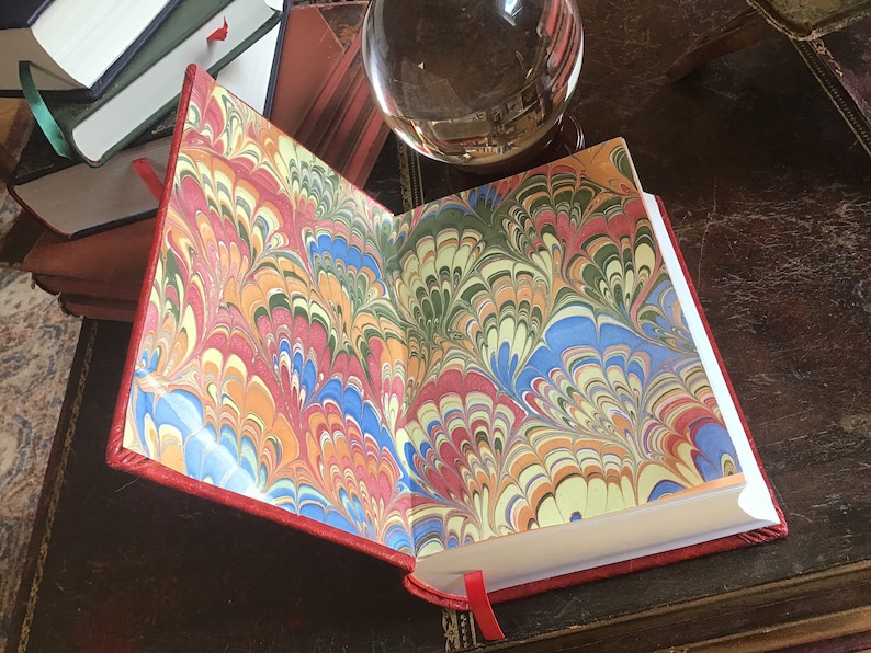 Puede incluir: Un diario encuadernado en cuero rojo con papeles de marfil marmoleados. El marmoleado presenta un patr&oacute;n de remolino de rojo, azul, verde, amarillo y blanco.