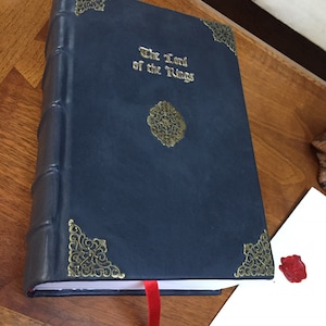 Puede incluir: Un libro encuadernado en cuero azul oscuro con detalles dorados y el t&iacute;tulo "El Se&ntilde;or de los Anillos" en relieve en la portada. Un marcap&aacute;ginas de cinta roja est&aacute; unido al libro.