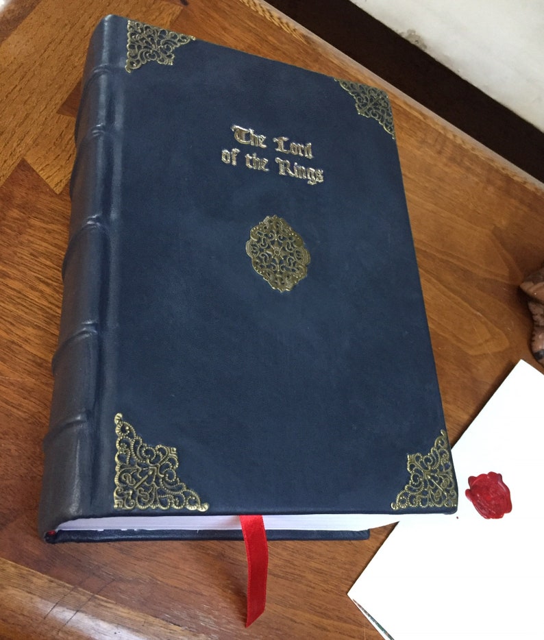 Puede incluir: Un libro encuadernado en cuero azul oscuro con detalles dorados y el t&iacute;tulo "El Se&ntilde;or de los Anillos" en relieve en la portada. Un marcap&aacute;ginas de cinta roja est&aacute; unido al libro.