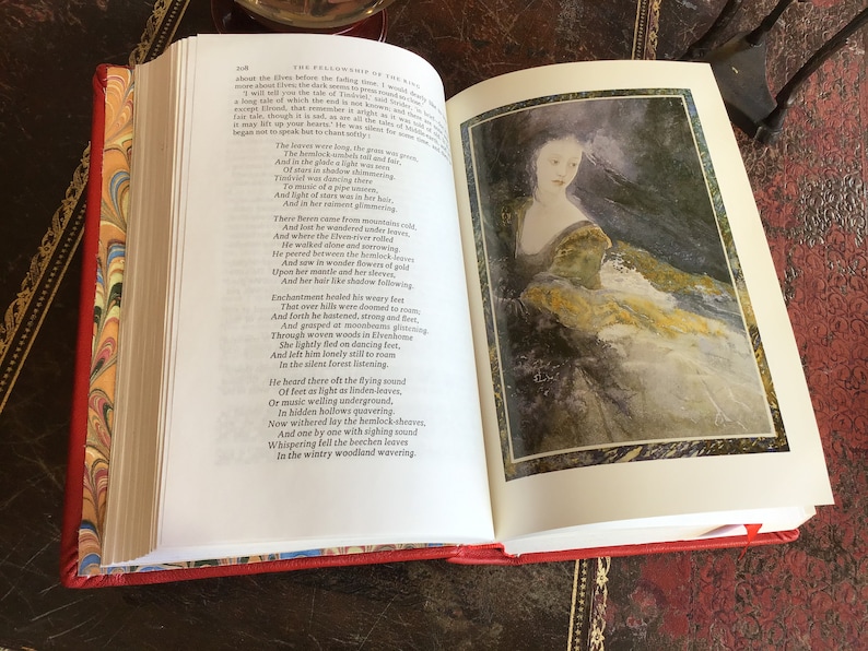 Puede incluir: Un libro abierto con una cubierta roja y p&aacute;ginas de guarda con vetas. El libro est&aacute; abierto en una p&aacute;gina con texto sobre los elfos y una ilustraci&oacute;n de acuarela de una mujer con un vestido fluido. El texto dice "La Comunidad del Anillo" e incluye un poema sobre los elfos.