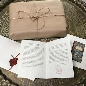 Puede incluir: Un paquete de papel marr&oacute;n atado con cuerda, una tarjeta blanca con el texto "The Bookbindery Custom Made Books" y una tarjeta peque&ntilde;a con una imagen de portada de libro. El texto en la tarjeta dice "Bookbinding at the University of New Mexico".