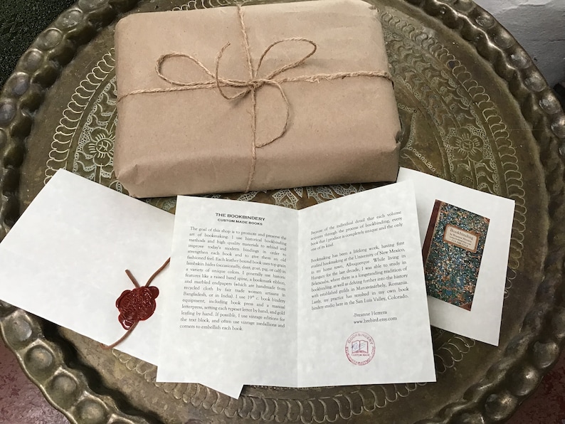 Puede incluir: Un paquete de papel marr&oacute;n atado con cuerda, una tarjeta blanca con el texto "The Bookbindery Custom Made Books" y una tarjeta peque&ntilde;a con una imagen de portada de libro. El texto en la tarjeta dice "Bookbinding at the University of New Mexico".