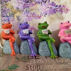 May include: Four colorful frog figurines, each holding a coffee cup. The frogs are orange, purple, green, and pink, sitting on gray rocks. Each rock has the text "Coffee gets me JUMPIN'". The coffee cups have the word "coffee".