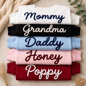 Pode incluir: Uma pilha de camisolas de gola redonda em branco, preto, azul claro, rosa e bordô. Cada camisola tem um nome bordado na frente: "Mommy", "Grandma", "Daddy", "Honey" e "Poppy".