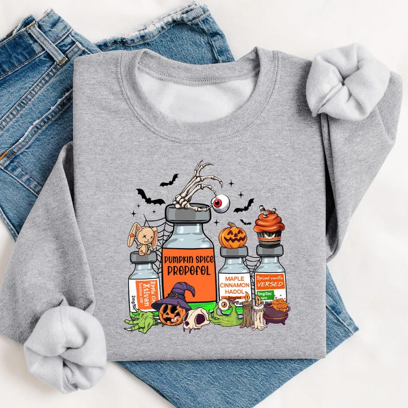 May include: A light grey sweatshirt with a Halloween graphic. The design features potion bottles labelled "Pumpkin Spice Propofol" and "Maple Cinnamon Hadol", alongside bats, a skeleton hand, and a jack-o'-lantern.