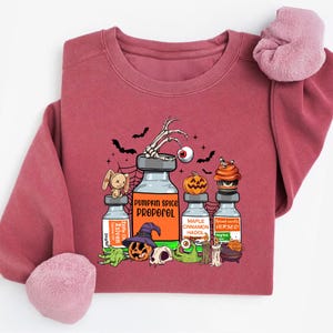 May include: A maroon sweatshirt with a Halloween graphic. The design features vials labelled "Pumpkin Spice Propofol", "Maple Cinnamon Hado", and "Spiced Vanilla Versed", alongside bats, a skeleton hand, and a pumpkin.