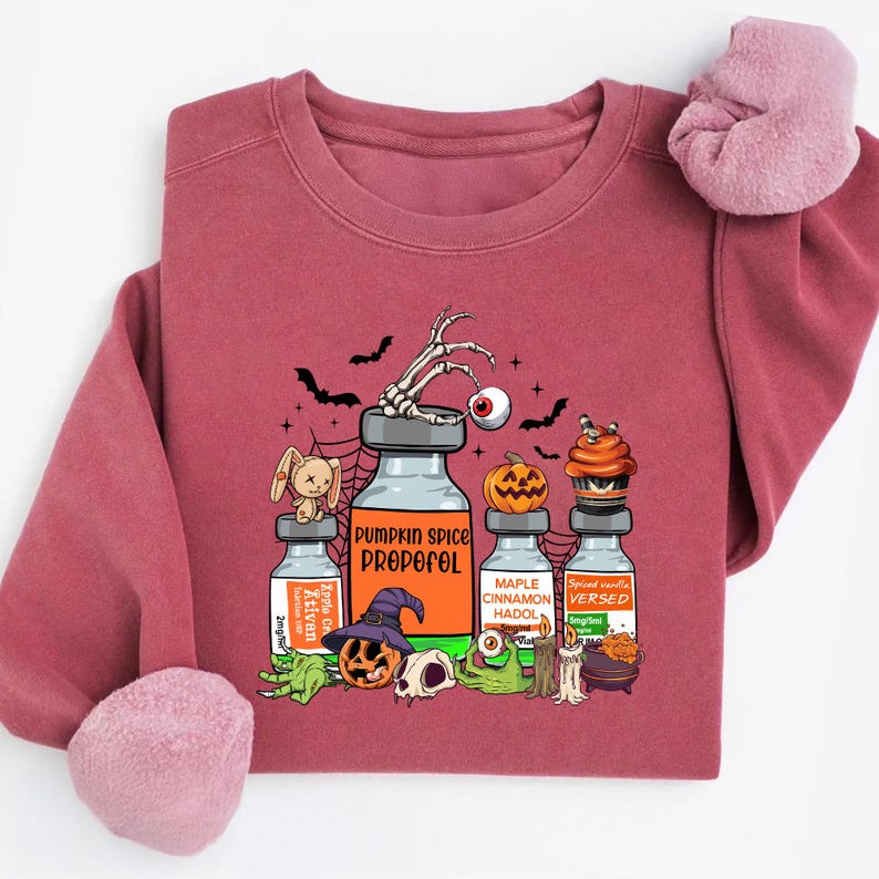 May include: A maroon sweatshirt with a Halloween graphic. The design features vials labelled "Pumpkin Spice Propofol", "Maple Cinnamon Hado", and "Spiced Vanilla Versed", alongside bats, a skeleton hand, and a pumpkin.
