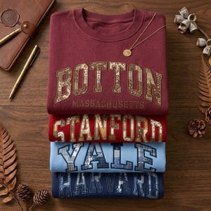 Pode incluir: Uma pilha de quatro camisolas dobradas em bordô, vermelho, azul claro e azul marinho. Cada camisola apresenta o nome de uma universidade: Boston, Stanford, Yale e Harvard. Um colar e uma caneta dourados são visíveis.