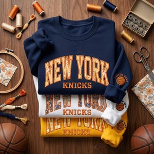 May include: A stack of three crewneck sweatshirts in navy blue, white, and yellow. Each features the text NEW YORK KNICKS in an arched font with a patterned fill. The sleeves display a basketball graphic with the text CUSTOM NAME.