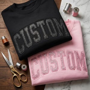 Puede incluir: Dos sudaderas, una negra y otra rosa, cada una bordada con la palabra "CUSTOM" en una fuente brillante de estilo universitario. Los útiles de costura, incluyendo tijeras y carretes de hilo, están dispuestos alrededor de las sudaderas.