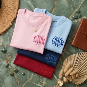 May include: Four crew neck t-shirts in pink, light blue, navy, and burgundy, each embroidered with a monogram. A gold necklace rests on the pink shirt. The shirts are stacked on a green marble surface, with a straw hat and a leather journal nearby.