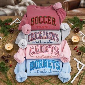 May include: Four crewneck sweatshirts in shades of pink, grey and blue, stacked on a wooden surface. Each sweatshirt has a different team name or title embroidered on the front. The top one says "Soccer Mom".