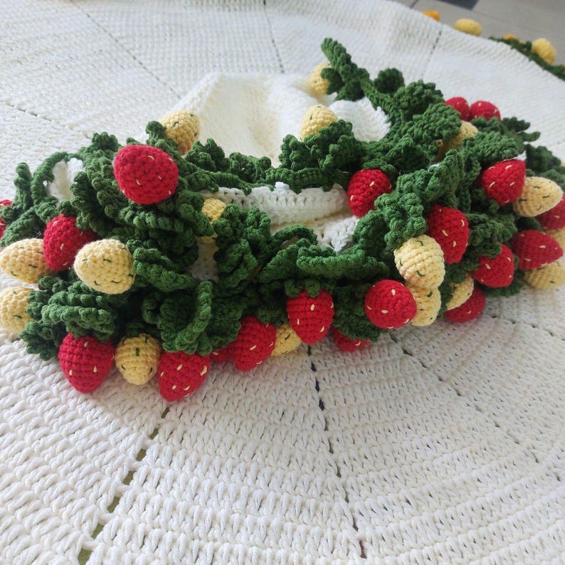 May include: A white crocheted blanket with a green and red crocheted strawberry garland. The garland is made of individual crocheted strawberries and leaves.