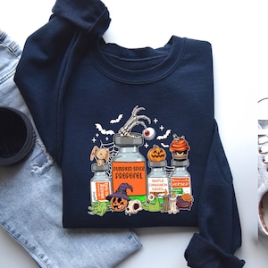 May include: Navy blue sweatshirt with a Halloween graphic. The design includes potion bottles labelled "Pumpkin Spice Propoful", "Ketivan", "Maple Cinnamon Hadol", and "Spiced Vanilla Versed". Features bats, a skeleton hand, and a jack-o'-lantern.