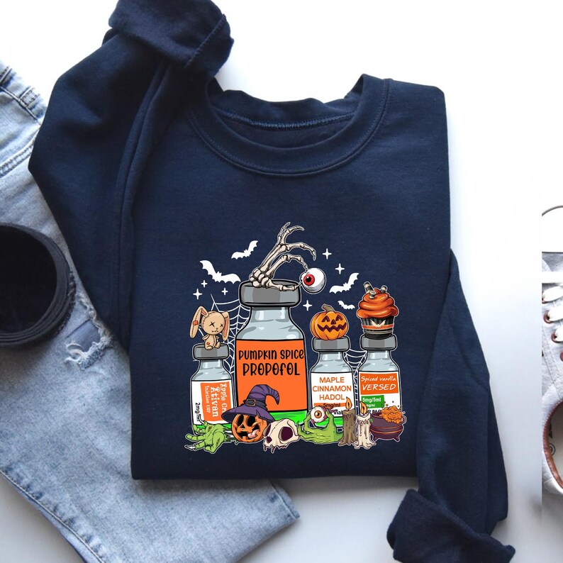 May include: Navy blue sweatshirt with a Halloween graphic. The design includes potion bottles labelled "Pumpkin Spice Propoful", "Ketivan", "Maple Cinnamon Hadol", and "Spiced Vanilla Versed". Features bats, a skeleton hand, and a jack-o'-lantern.