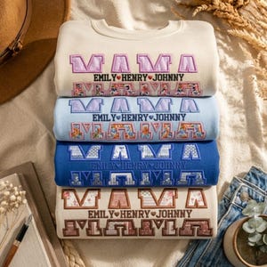 May include: A stack of four sweatshirts in cream, light blue, and navy blue. Each sweatshirt has the embroidered word "MAMA" and the names "EMILY HENRY JOHNNY". The bottom sweatshirt is a cream color with a brown and pink floral pattern.