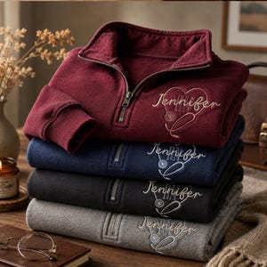 Könnte beinhalten: Ein Stapel aus vier Sweatshirts mit kurzem Reißverschluss in Kastanienbraun, Marineblau, Schwarz und Grau. Jedes Sweatshirt ist auf der Brust mit einem Herz, einem Stethoskop, den Buchstaben RN und dem Namen Jennifer in weißer Schreibschrift bestickt.