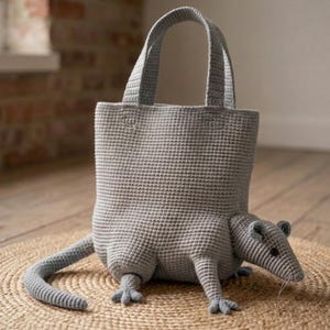 Könnte beinhalten: Eine graue, gehäkelte Tragetasche in Rattenform mit einem langen Schwanz, vier kleinen Beinen, einer spitzen Schnauze und zwei kleinen Ohren. Die Tasche hat einen rechteckigen Körper mit flachem Boden und zwei stabilen Tragegriffen an der Oberseite.