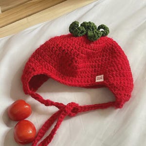 Pode incluir: Chapéu vermelho de crochê em forma de morango com folhas verdes de crochê no topo. O chapéu tem uma longa gravata que pode ser amarrada sob o queixo. O chapéu tem uma pequena etiqueta que diz "feito à mão".