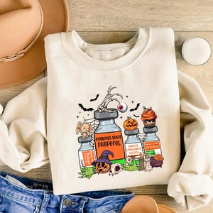 May include: A cream-coloured sweatshirt with a Halloween graphic. The design features cartoon potion bottles labelled "Pumpkin Spice Propofol" and "Maple Cinnamon Hadol", alongside bats, a skeleton hand, and a jack-o'-lantern.