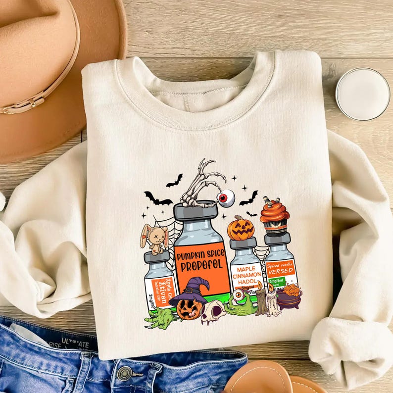 May include: A cream-coloured sweatshirt with a Halloween graphic. The design features cartoon potion bottles labelled "Pumpkin Spice Propofol" and "Maple Cinnamon Hadol", alongside bats, a skeleton hand, and a jack-o'-lantern.