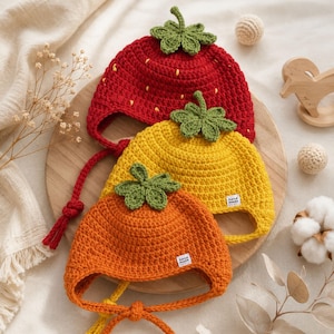 May include: Three handmade crochet hats featuring fruit-inspired designs. The top hat is red with yellow seed accents, the middle is yellow, and the bottom is orange. Each hat includes a green leaf topper and chin ties with a small white tag.