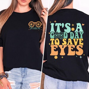 Puede incluir: Camiseta negra con un gráfico de estilo retro en la espalda que dice "It's a good day to save eyes". La parte delantera de la camiseta tiene un par de gafas con el nombre "Jessica" escrito debajo.
