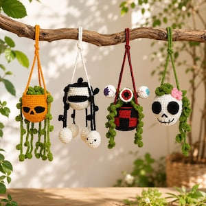 Puede incluir: Cuatro macetas colgantes de ganchillo suspendidas de una rama. La primera es una calavera naranja con enredaderas verdes, la segunda un fantasma blanco y negro con esferas colgantes, la tercera un macetero rojo y negro con ojos y la cuarta una calavera.