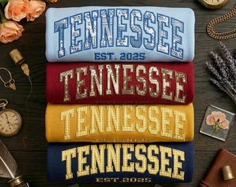 Puede incluir: Cuatro sudaderas apiladas en azul claro, burdeos, amarillo y azul marino, cada una bordada con "TENNESSEE" en una fuente universitaria y "EST. 2025". Las sudaderas están dobladas y dispuestas sobre una superficie de madera oscura, con accesorios vintage y flores.