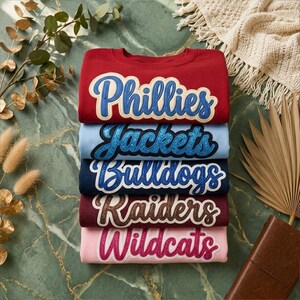 May include: A stack of five crewneck sweatshirts in various colors: red, light blue, navy, burgundy, and pink. Each sweatshirt features a team name in a script font with a contrasting outline. The names are "Phillies", "Jackets", "Bulldogs", "Raiders", and "Wildcats".
