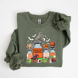 May include: An olive green sweatshirt with a Halloween graphic. The graphic includes cartoon vials labelled "Pumpkin Spice Propofol," "Maple Cinnamon Hadol," and "Spiced Vanilla Versed." Other elements include a skeleton hand, pumpkins, and a cupcake.
