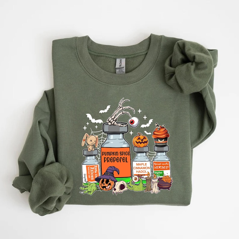 May include: An olive green sweatshirt with a Halloween graphic. The graphic includes cartoon vials labelled "Pumpkin Spice Propofol," "Maple Cinnamon Hadol," and "Spiced Vanilla Versed." Other elements include a skeleton hand, pumpkins, and a cupcake.