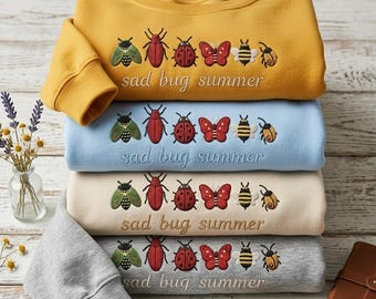 May include: A stack of four sweatshirts in mustard yellow, light blue, beige, and gray. Each sweatshirt features an embroidered design of various insects and the text "sad bug summer".