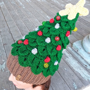 May include: A green crocheted Christmas tree hat with red and white ornaments and yellow lights. The hat is worn on a brown mannequin head.