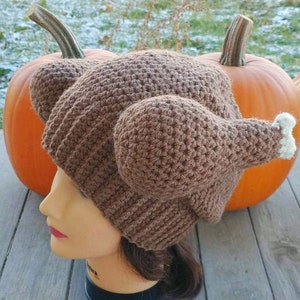 May include: A brown crocheted hat shaped like a turkey with white accents on the wings. The hat is worn on a mannequin head with brown hair.