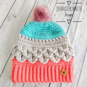 May include: A crocheted hat with a pink pom pom. The hat is made in three colors: teal, beige, and coral. The hat has a decorative flower embellishment.