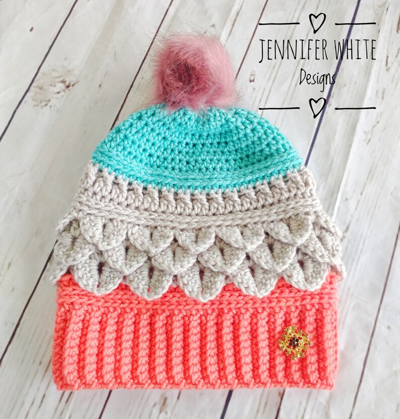 May include: A crocheted hat with a pink pom pom. The hat is made in three colors: teal, beige, and coral. The hat has a decorative flower embellishment.