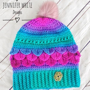 May include: A colorful crocheted hat with a pink pom pom on top. The hat has a teal, purple, pink, and blue color gradient. The hat has a decorative green and gold embellishment on the side. The hat is made by Jennifer White Designs.