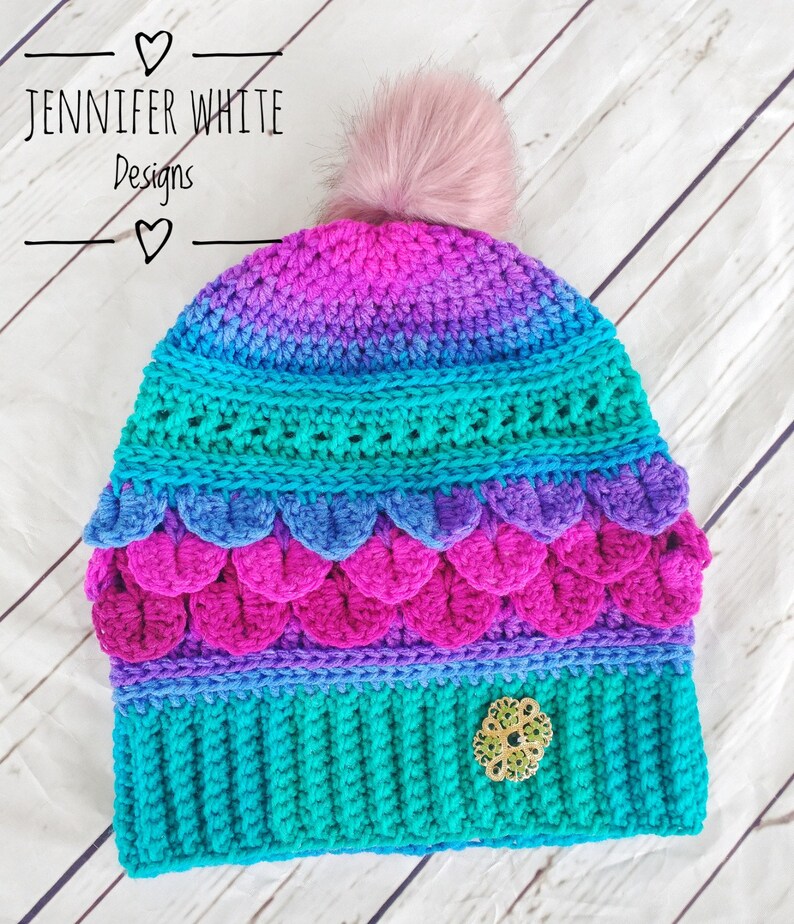 May include: A colorful crocheted hat with a pink pom pom on top. The hat has a teal, purple, pink, and blue color gradient. The hat has a decorative green and gold embellishment on the side. The hat is made by Jennifer White Designs.