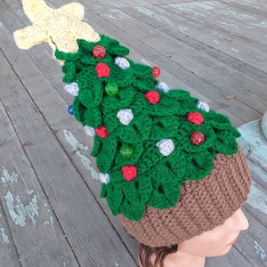 May include: A green crocheted Christmas tree hat with a yellow star on top. The hat is decorated with red, white, and blue ornaments. The hat is worn on a mannequin head.
