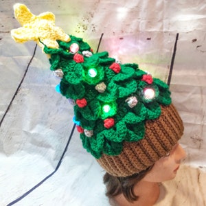 May include: A green crocheted Christmas tree hat with a yellow star on top. The hat has red and silver ornaments and lights.