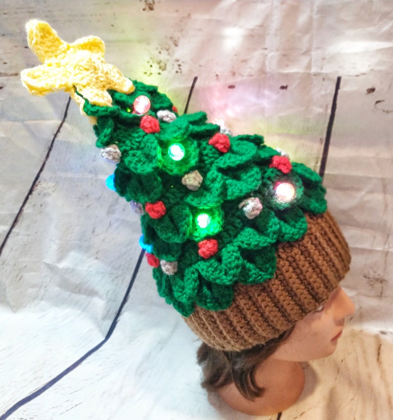 May include: A green crocheted Christmas tree hat with a yellow star on top. The hat has red and silver ornaments and lights.