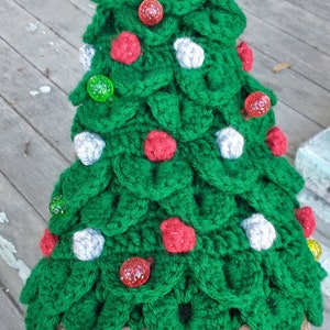 May include: A green crocheted Christmas tree hat with a yellow star on top. The hat is decorated with red, green, and white ornaments. The hat is brown on the bottom and has a tag that says "Hat"