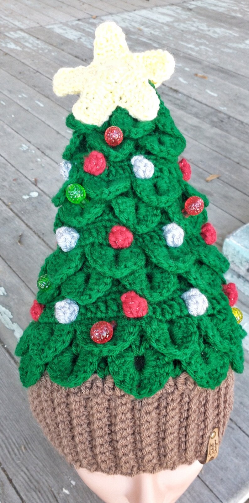 May include: A green crocheted Christmas tree hat with a yellow star on top. The hat is decorated with red, green, and white ornaments. The hat is brown on the bottom and has a tag that says "Hat"